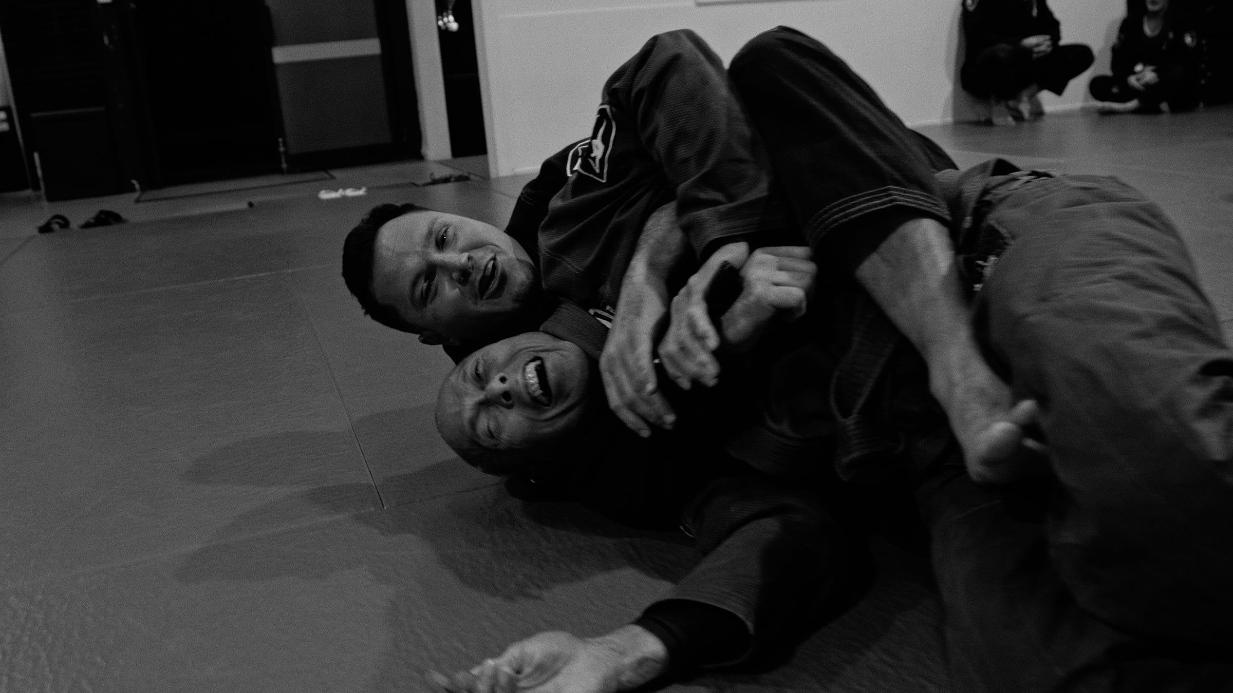 Black and white photo of two people grappling on a mat in an indoor training space, with a third person seated in the background.