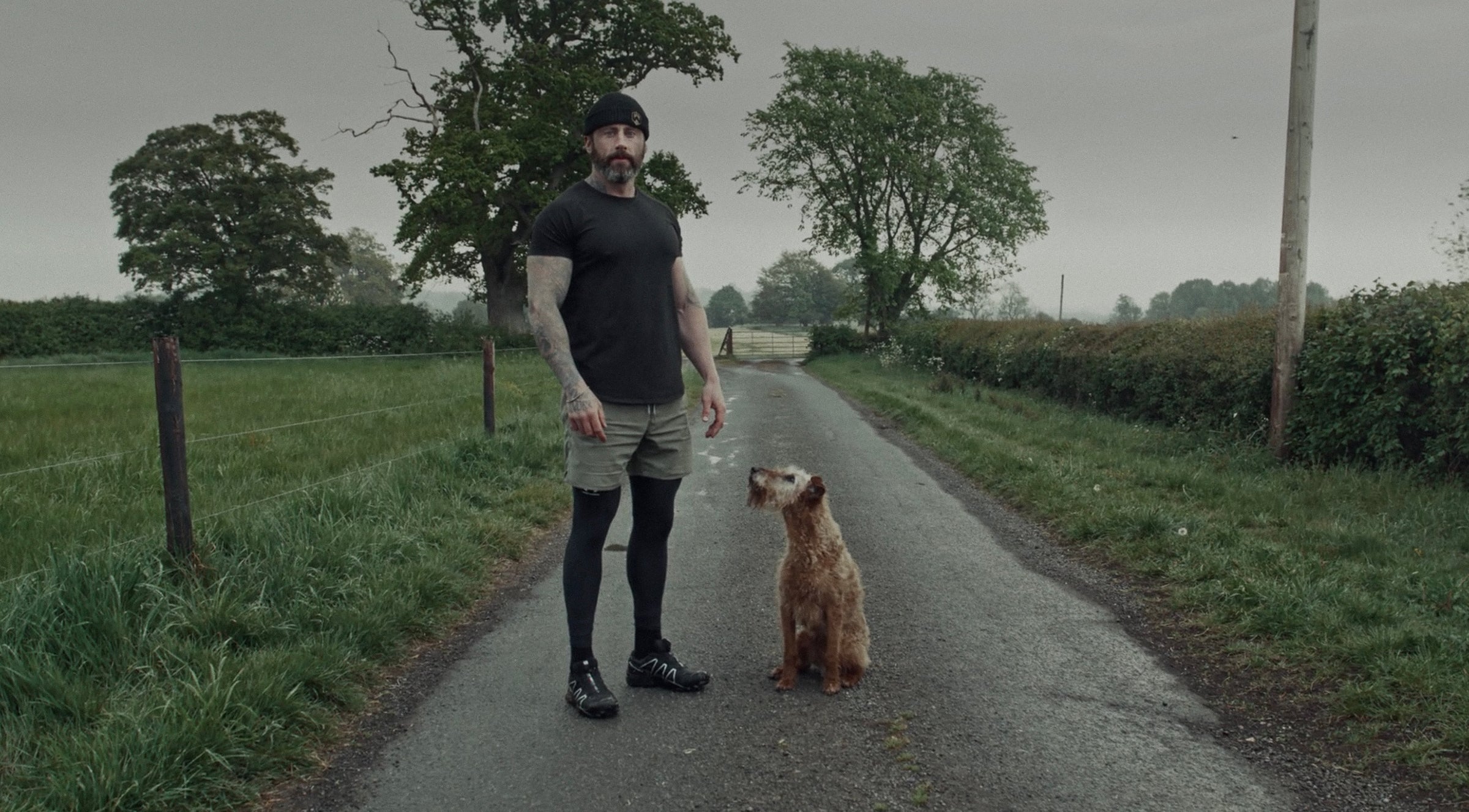Man standing on a rural road with a dog, surrounded by greenery.