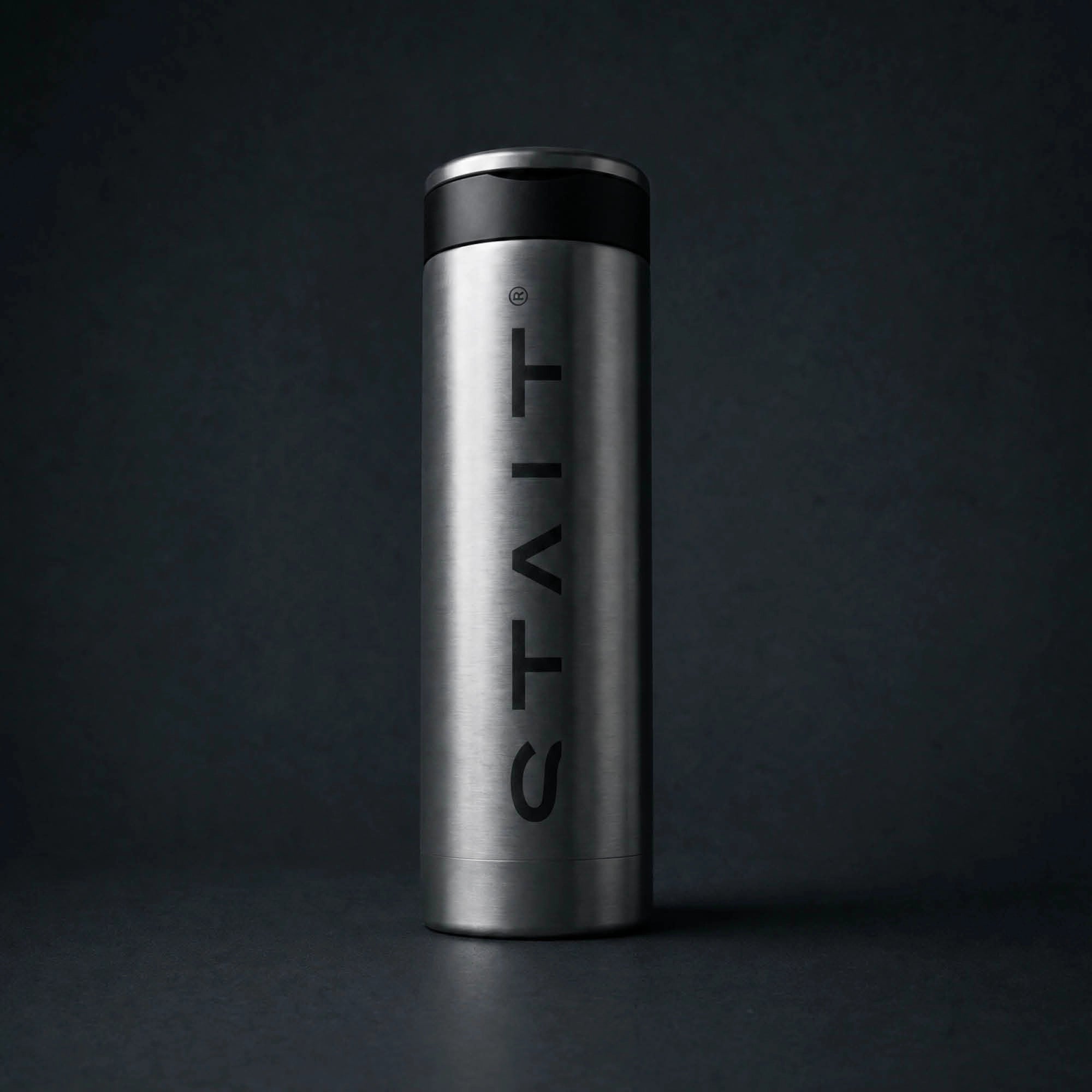A stainless steel travel tumbler with a black lid and the brand name 'STAIT' printed vertically in black on the front, set against a dark gray background.