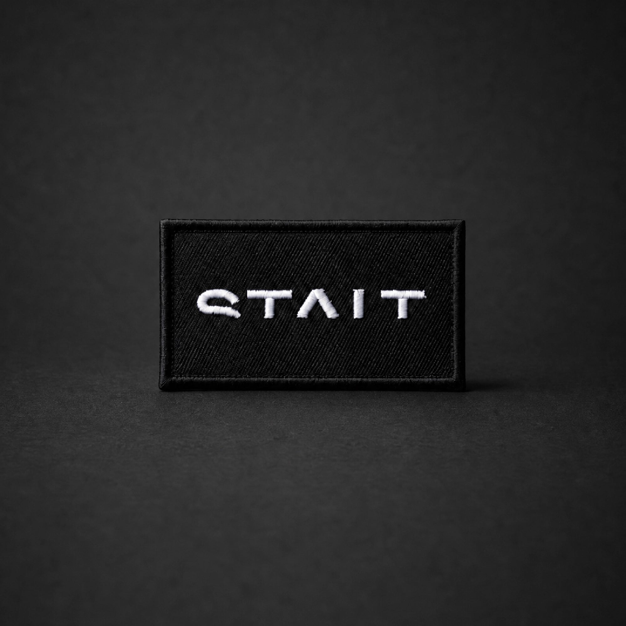 A rectangular black embroidered patch with the word 'STAIT' in white stylized lettering, centered on a dark gray background.