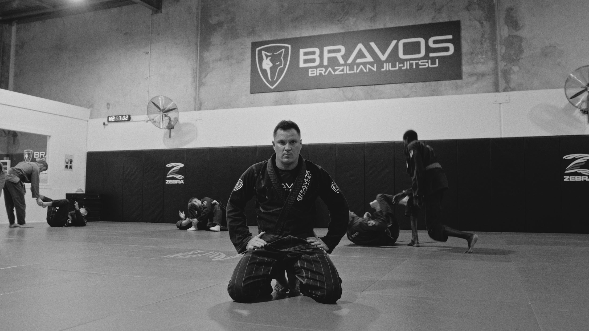 Rodrigo Giraldes in a black Brazilian Jiu-Jitsu gi kneels on a mat inside a gym, with a large 'BRAVO Brazilian Jiu-Jitsu' banner on the wall behind him and other people visible in the background.